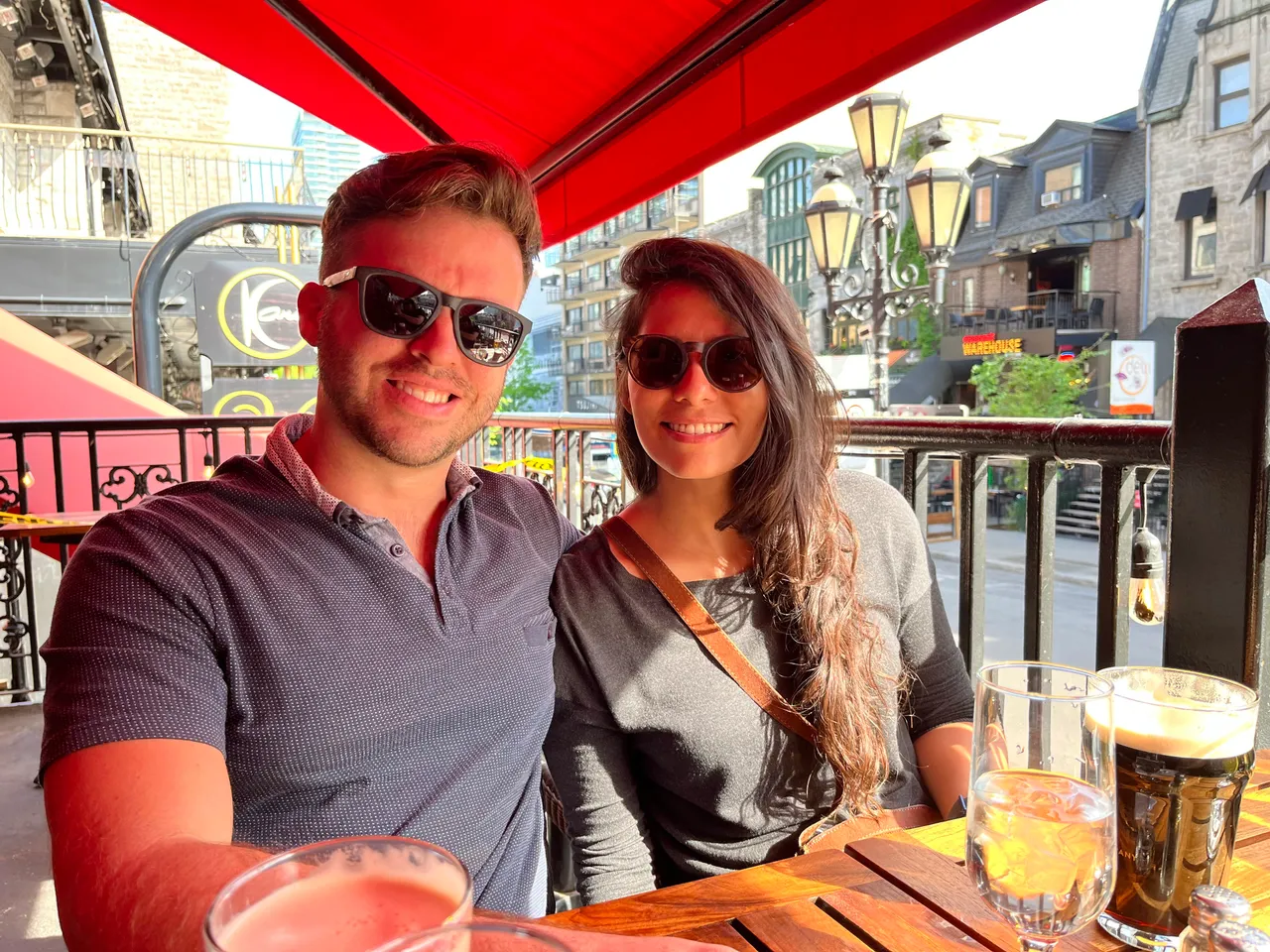 Patrick and his wife wearing sunglasses at an outdoor cafe under a red canopy.