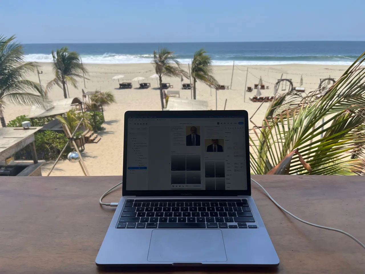 A laptop on a wooden desk overlooking a beach with palm trees.