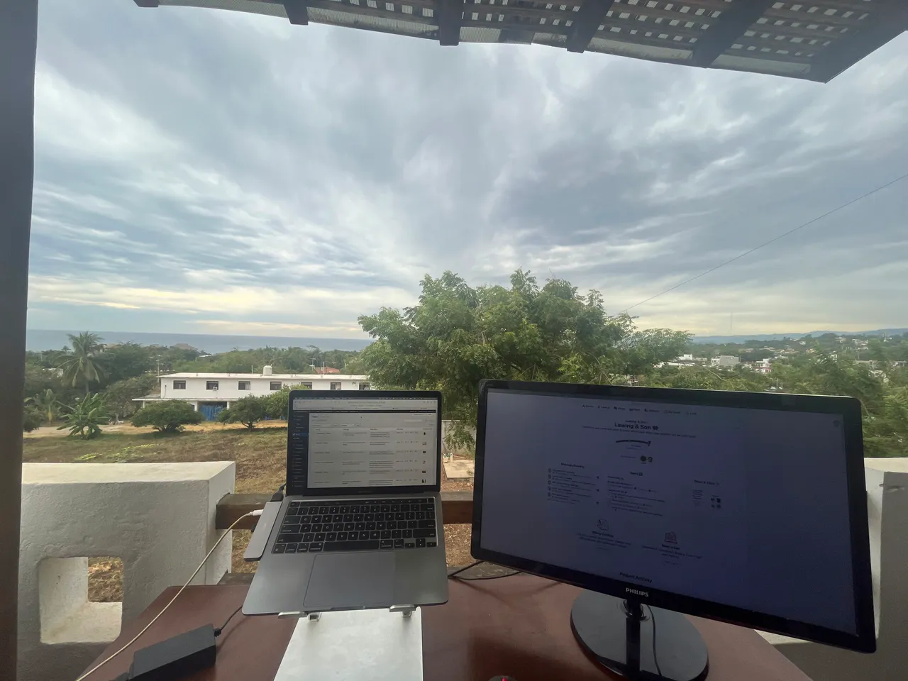 An outdoor workspace with a laptop and a monitor on a desk, green landscape and cloudy sky behind.