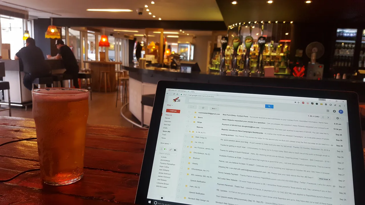 A laptop on a wooden bar table next to a glass of beer, with the pub's taps in the background.